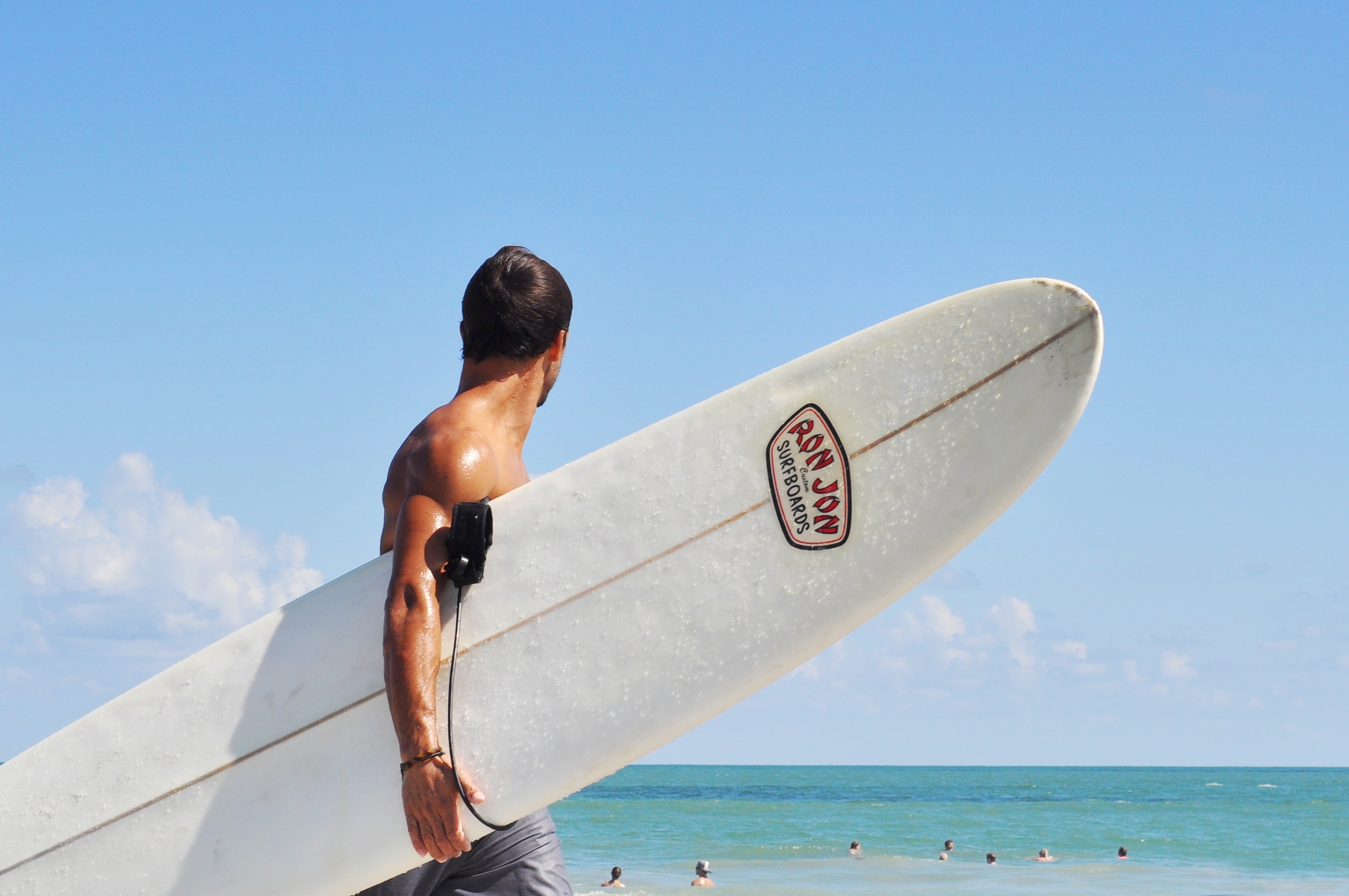 2896x1924 topless man carrying white ron jon surfboard and looking towards people  swimming on beach under blue sky