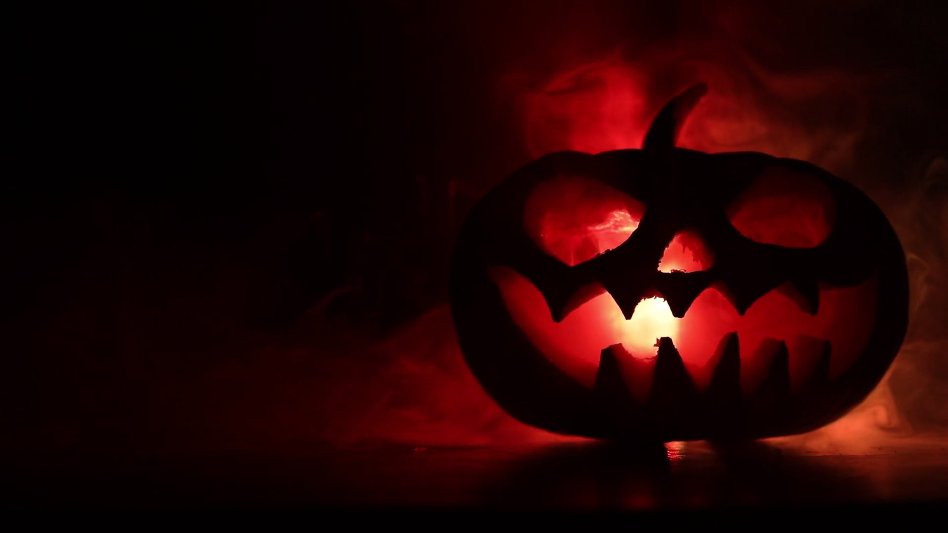 1920x1080 Scary Halloween pumpkin with carved eyes and a smile with burning candles  inside on a dark background with fire and fog. Empty space.